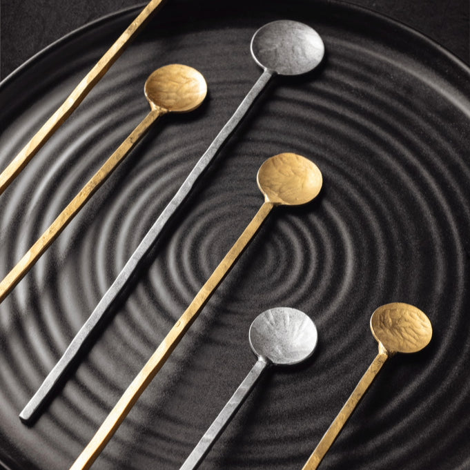 Small Silver and gold Catalina Stirring Spoons on a black plate.
