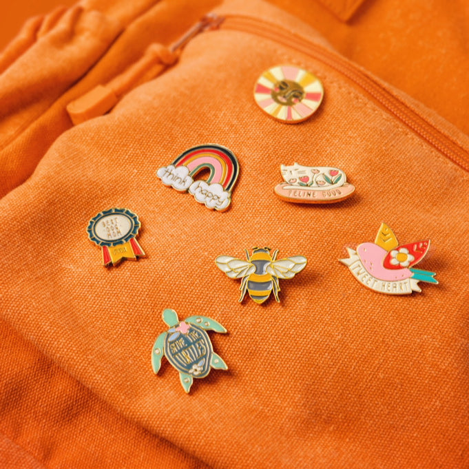 Rainbow, sunshine, cat, bird, bee, dog, and turtle enamel pins on an orange bag.