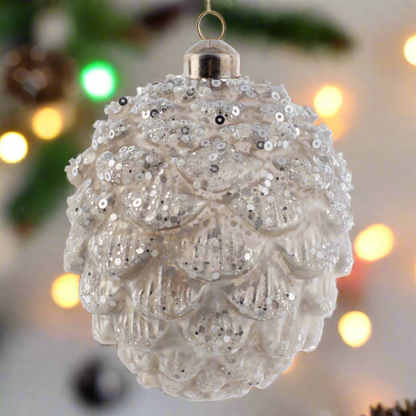 Small Glittered Glass Pinecone White Ornament with holiday lighting in the background. 