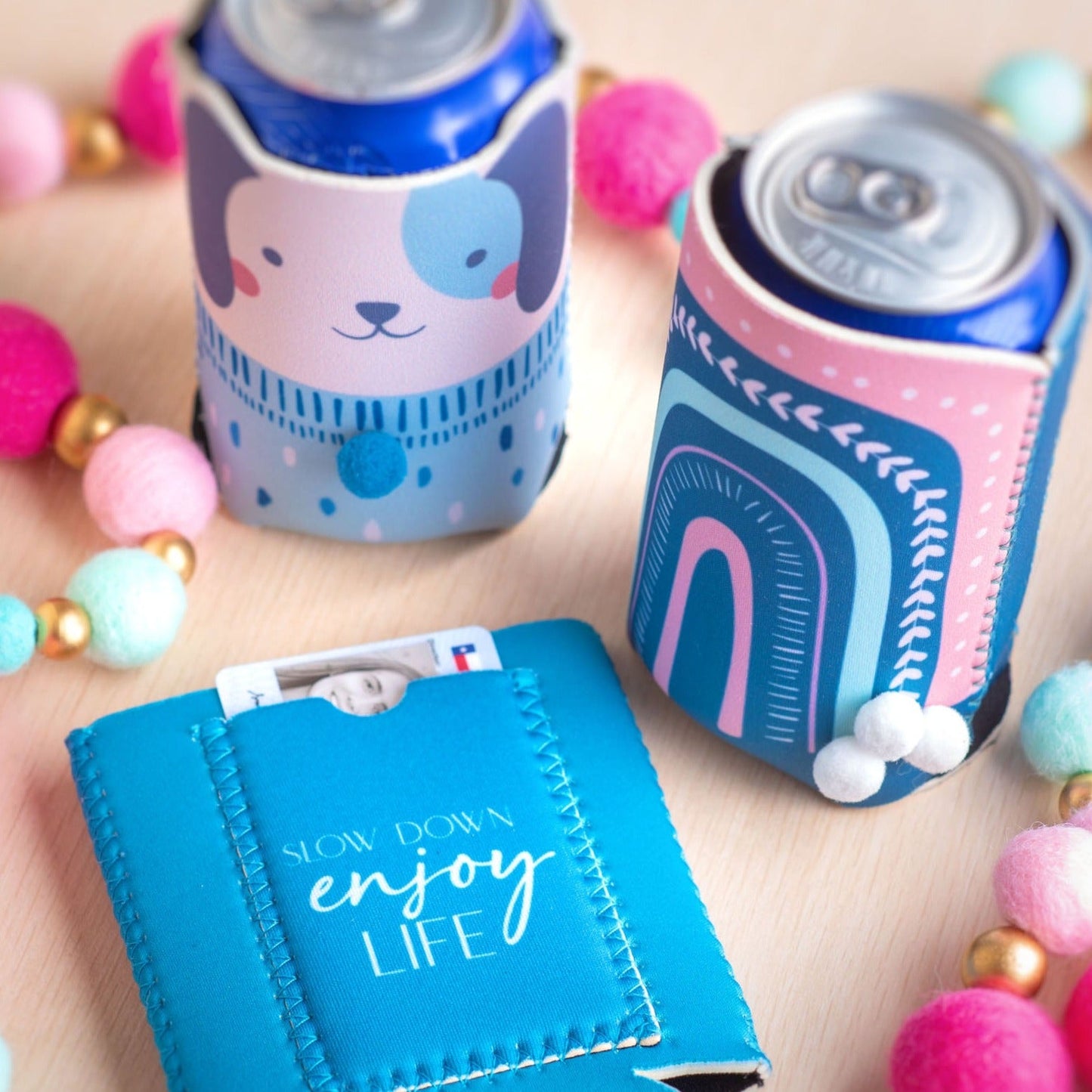 Dog, rainbow, and turtle can coolers on a table with cans and ID's in the pouch. 