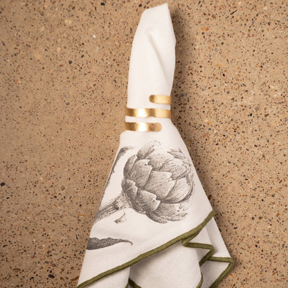Artichoke Merrowed Edge Dinner Napkins on a counter with gold napkin ring. 