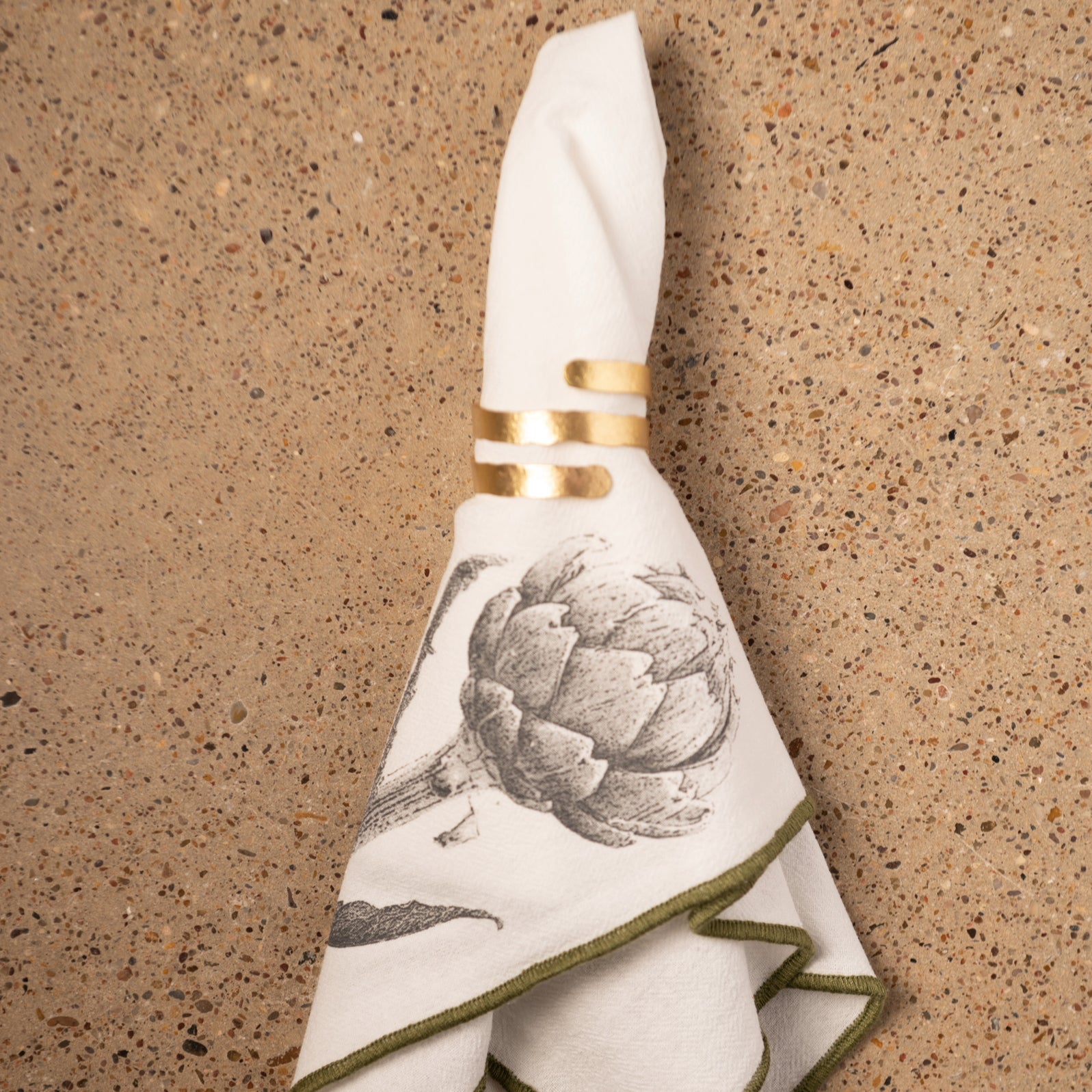 Artichoke Merrowed Edge Dinner Napkins on a counter with gold napkin ring. 